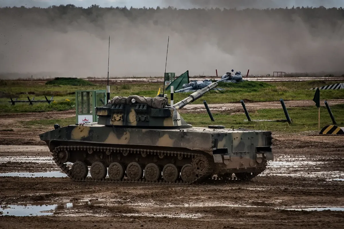 A military tank moves through a muddy training ground with dust in the background.
